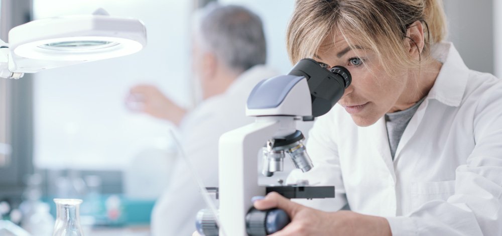 image of a midical lab with a female scientist looking into a microscope with another scientist on the background